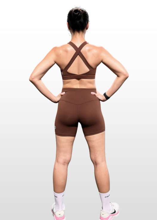 A woman shown from the back wearing a matching brown athletic set consisting of a strappy sports bra and high-waisted biker shorts, standing against a light gray gradient background.