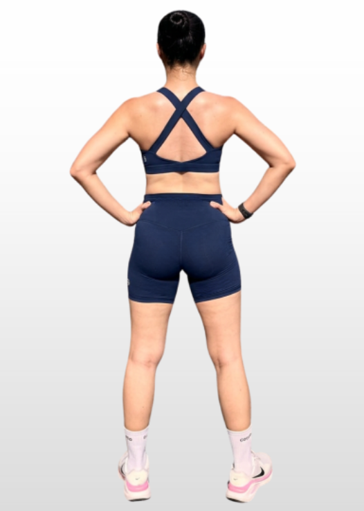A woman shown from the back wearing a navy blue sports bra and matching athletic shorts, standing against a light gray gradient background.