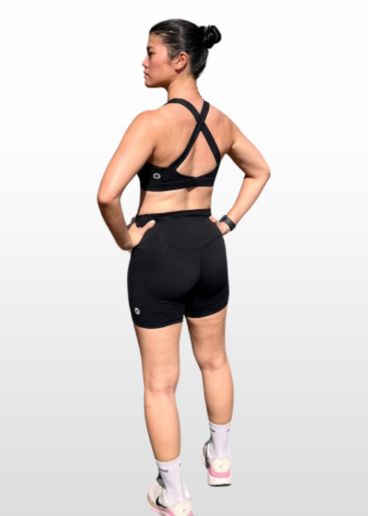 A woman shown from the back wearing a black sports bra and black athletic shorts, standing against a light gray gradient studio background.