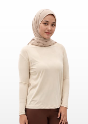 A woman modeling a beige long-sleeve top and brown flared pants with a matching beige hijab, standing against a light gray gradient background.