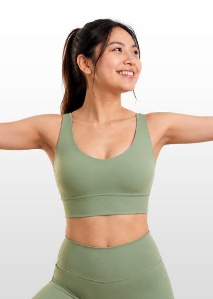 A woman with her hair in a ponytail is wearing a matching sage green sports bra and high-waisted leggings set, posing with her arms outstretched against a light gray gradient background.