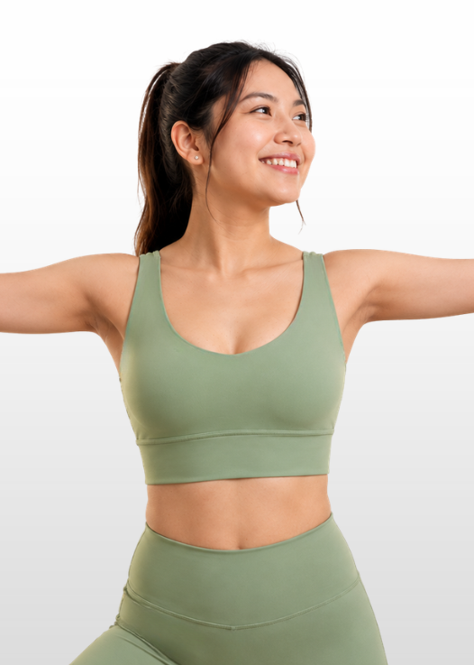 A woman with her hair in a ponytail is wearing a matching sage green sports bra and high-waisted leggings set, posing with her arms outstretched against a light gray gradient background.