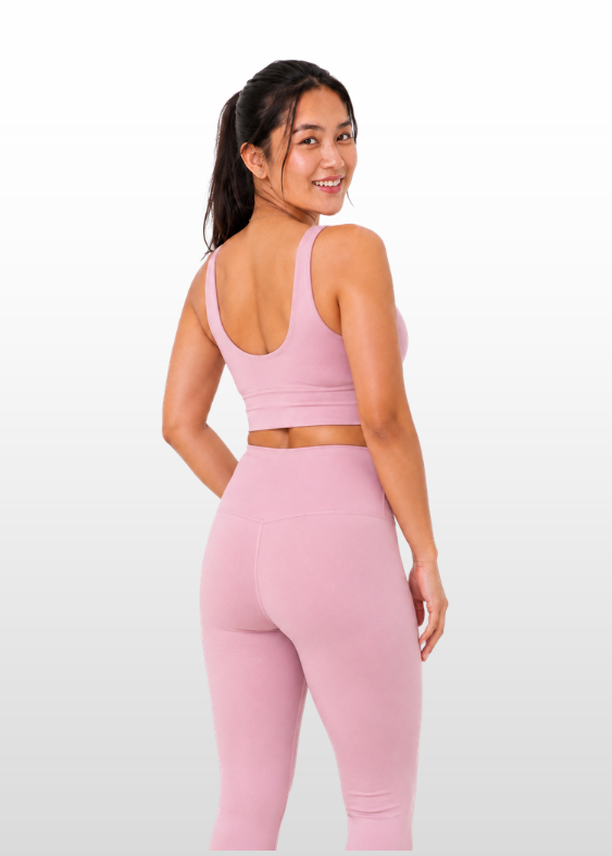A woman modeling a light pink activewear set consisting of a sports bra and high-waisted leggings, shown from a three-quarters back view against a light gray gradient background.