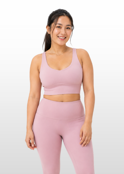 A woman modeling a matching dusty pink activewear set consisting of a scoop-neck sports bra and high-waisted leggings, set against a clean white studio background.
