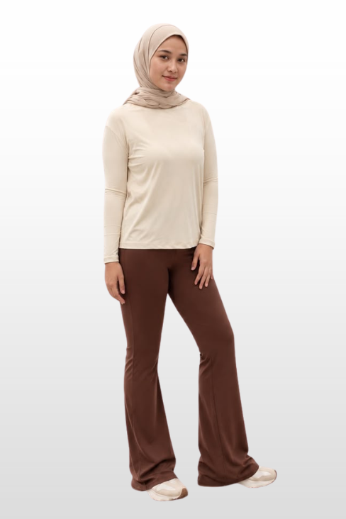 A woman modeling a beige long-sleeve top and brown flared pants with a matching beige hijab, standing against a light gray gradient background.