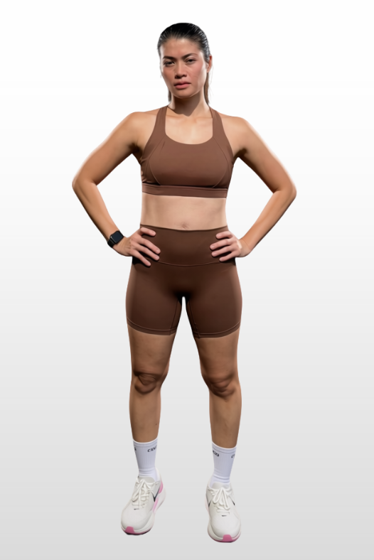 A woman modeling a matching brown activewear set consisting of a sports bra and high-waisted shorts, paired with white socks and white sneakers, set against a light gray gradient background.