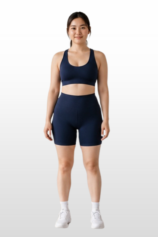 A woman stands facing forward wearing a matching navy blue sports bra and high-waisted athletic shorts, paired with white socks and sneakers, against a light gray gradient background.