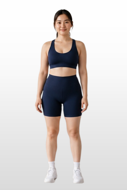 A woman stands facing forward wearing a matching navy blue sports bra and high-waisted athletic shorts, paired with white socks and sneakers, against a light gray gradient background.