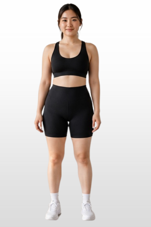 A woman modeling a matching black activewear set consisting of a sports bra and high-waisted shorts, paired with white sneakers, against a light gray gradient background.