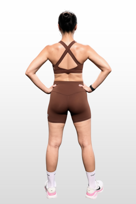 A woman shown from the back wearing a matching brown athletic set consisting of a strappy sports bra and high-waisted biker shorts, standing against a light gray gradient background.