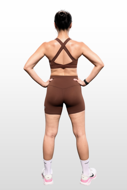 A woman shown from the back wearing a matching brown athletic set consisting of a strappy sports bra and high-waisted biker shorts, standing against a light gray gradient background.