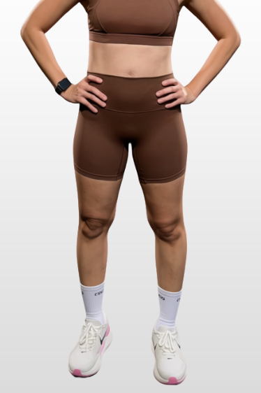 A woman modeling a matching brown activewear set consisting of a sports bra and high-waisted shorts, paired with white socks and white sneakers, set against a light gray gradient background.