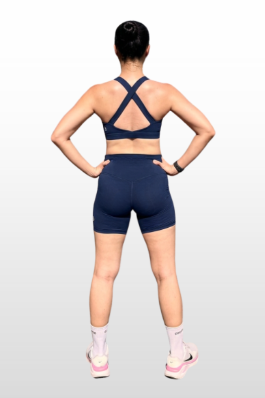 A woman shown from the back wearing a navy blue sports bra and matching athletic shorts, standing against a light gray gradient background.