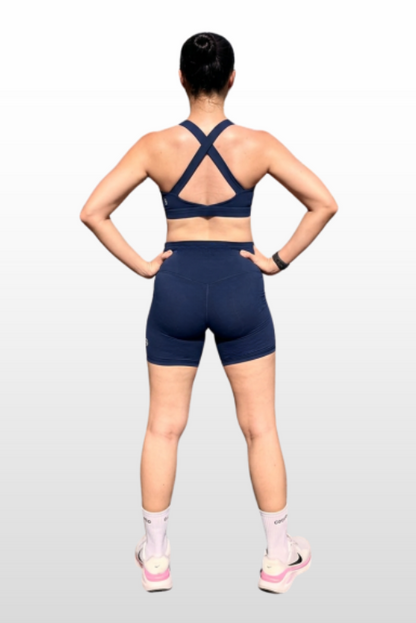 A woman shown from the back wearing a navy blue sports bra and matching athletic shorts, standing against a light gray gradient background.