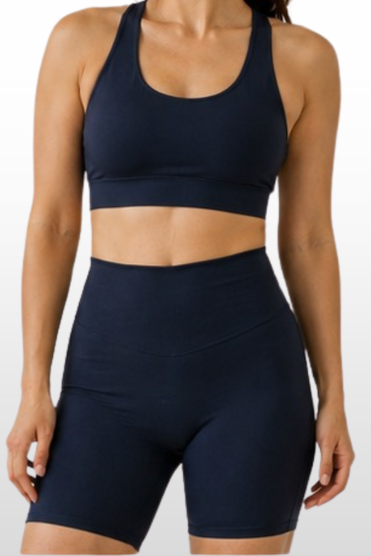 A woman modeling a matching navy blue activewear set consisting of a scoop-neck sports bra and high-waisted biker shorts, shown against a light gray gradient background.