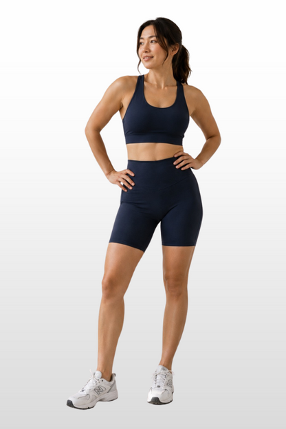 A woman modeling a matching navy blue sports bra and biker shorts activewear set, paired with white athletic sneakers, against a light gray gradient studio background.