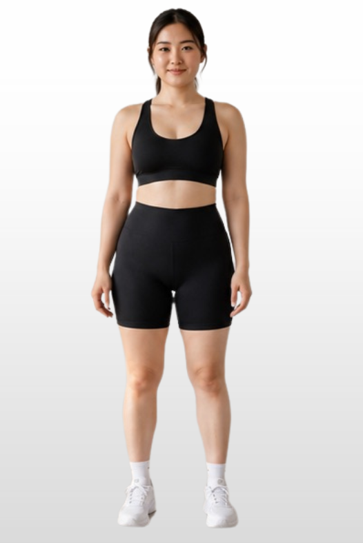 A woman modeling a matching black activewear set consisting of a sports bra and high-waisted shorts, paired with white sneakers, against a light gray gradient background.