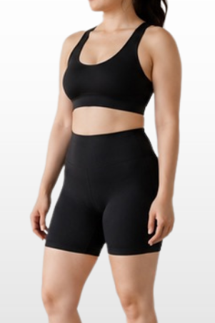 A woman modeling a black sports bra and matching high-waisted athletic shorts against a light gray gradient background.