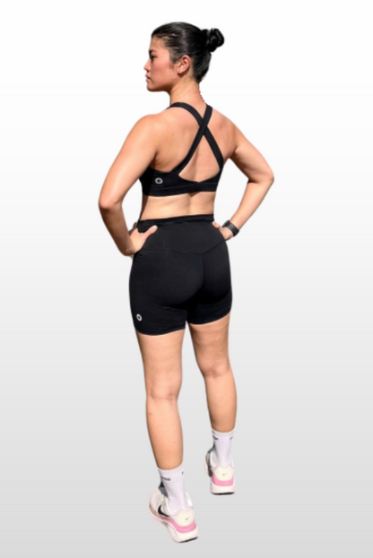 A woman shown from the back wearing a black sports bra and black athletic shorts, standing against a light gray gradient studio background.