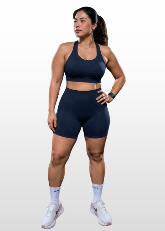A woman modeling a matching navy blue athletic set consisting of a sports bra and high-waisted shorts, paired with white athletic socks and sneakers, against a light gray gradient background.