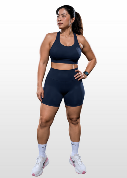 A woman modeling a matching navy blue athletic set consisting of a sports bra and high-waisted shorts, paired with white athletic socks and sneakers, against a light gray gradient background.