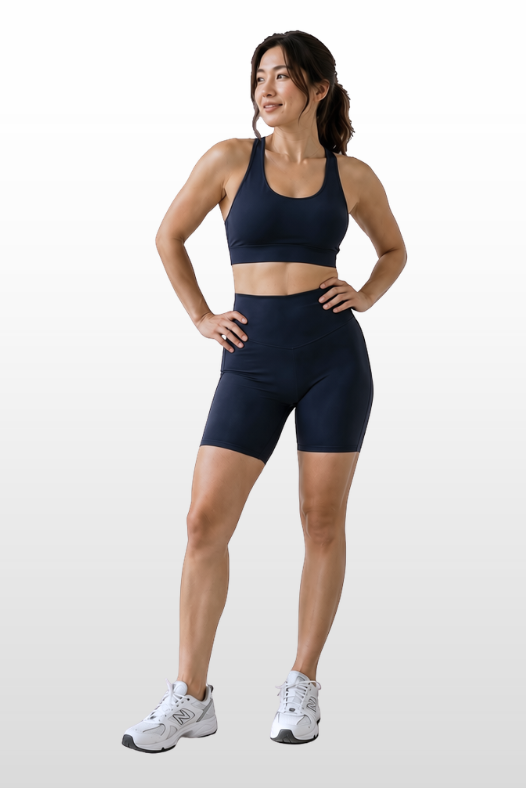 A woman modeling a matching navy blue sports bra and biker shorts activewear set, paired with white athletic sneakers, against a light gray gradient studio background.
